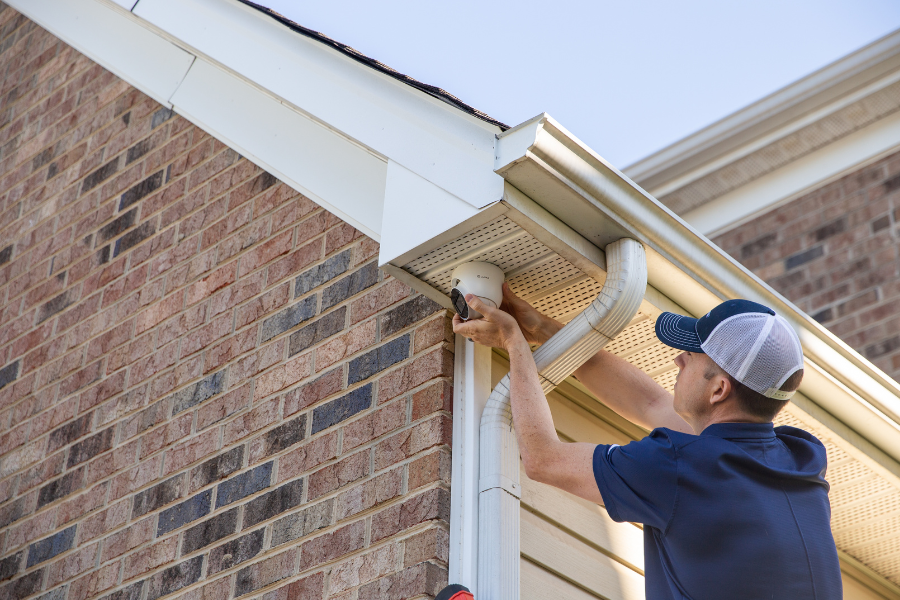 Technician installing an outdoor security camera under a roof eave on a brick home, highlighting professional surveillance installation for reliable exterior monitoring and protection.