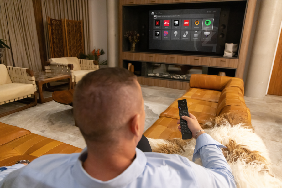 Person relaxing on a couch using a remote to control a wall-mounted smart home display, highlighting easy, centralized control of entertainment and home automation systems.