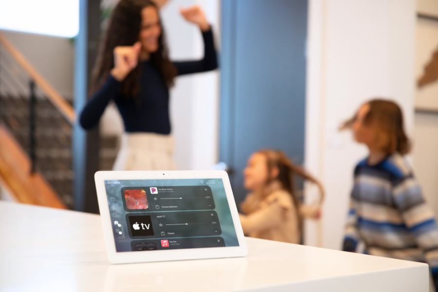 Smart home touchscreen control panel on a kitchen counter with a family in the background, highlighting easy, centralized control of entertainment and connected home systems.