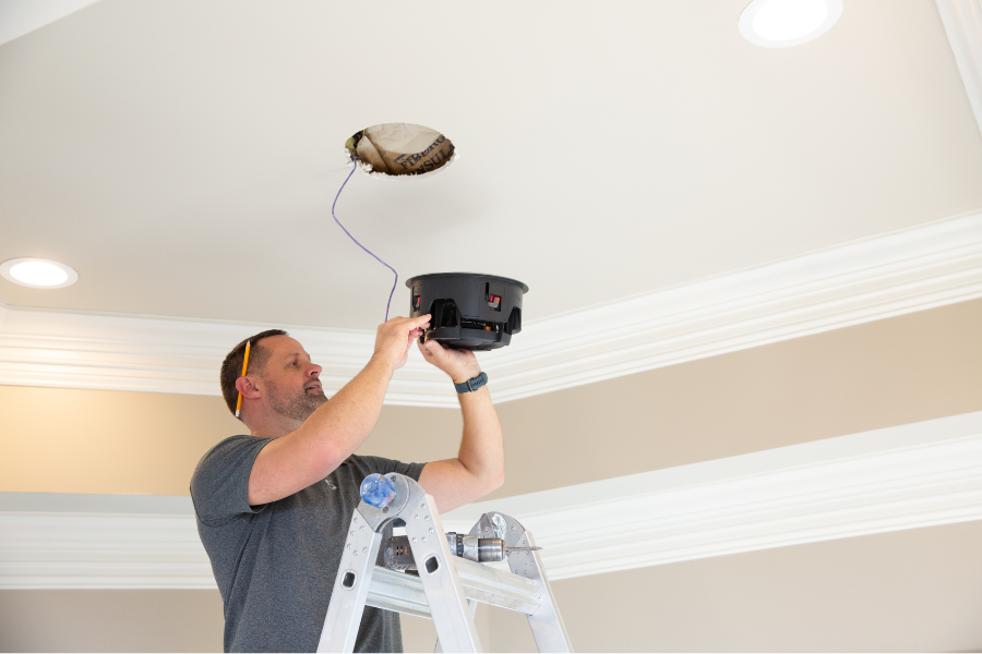 Technician on a ladder installing a ceiling-mounted smart device or sensor, highlighting professional setup of home security and low-voltage systems.